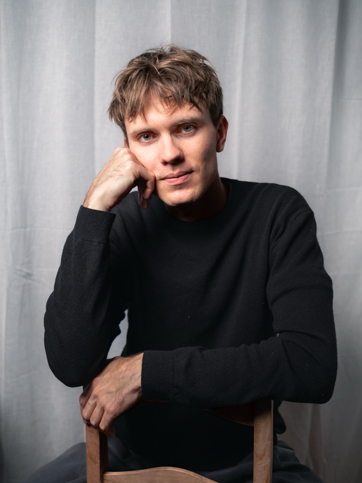 Jannes Hölzel, Fotograf von lumo.studios, sitzt rückwärts auf einem Holzstuhl vor einem hellen Vorhang